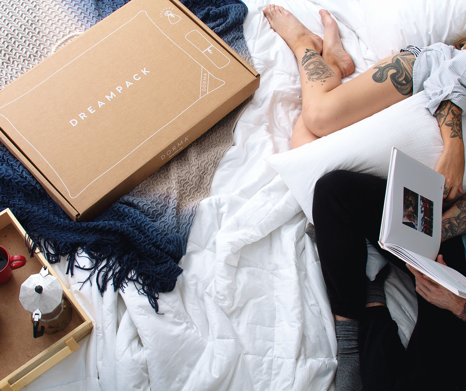 En la imagen se muestra una pareja sobre su cama leyendo un libro y en costado una frazada con degradados de azul y beige y sobre ella una caja de dreampack. 