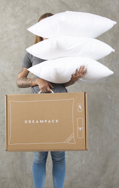 en un fondo gris se muestra una mujer sosteniendo 3 almohadas de forma apilada en la mano izquierda, y en la derecha una caja de dreampack.